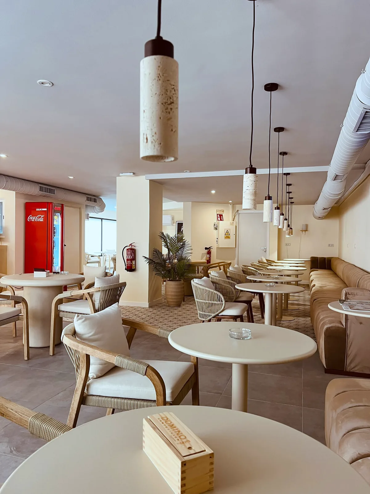 Tables and armchairs under pendant lamps, with plants and a long velvet bench